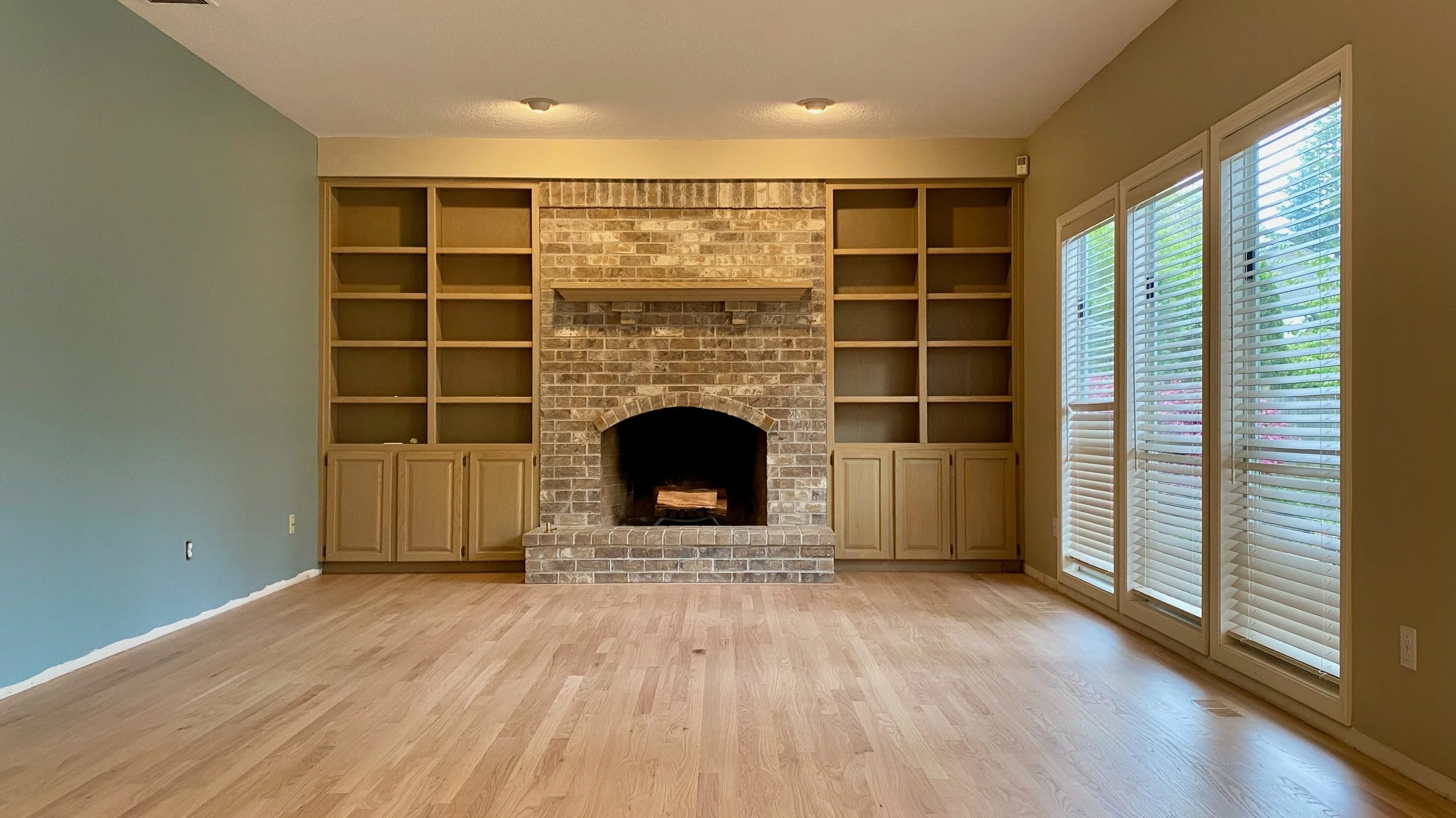 Empty living room with built-in wooden shelves and a brick fireplace, large windows with blinds, and hardwood flooring.