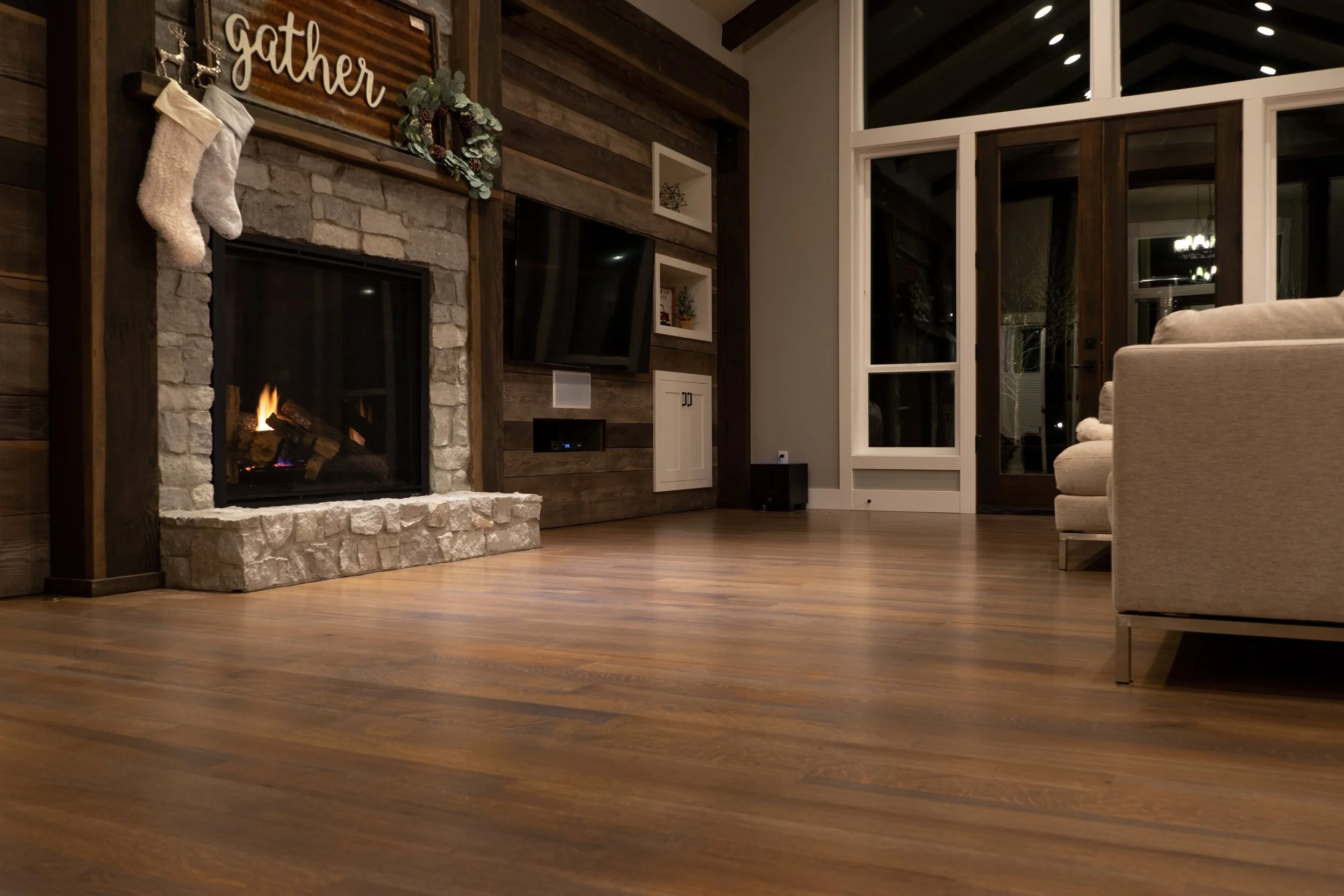 Living room with a wooden floor, a stone fireplace with holiday stockings, a wall-mounted TV, and large glass doors leading outside.