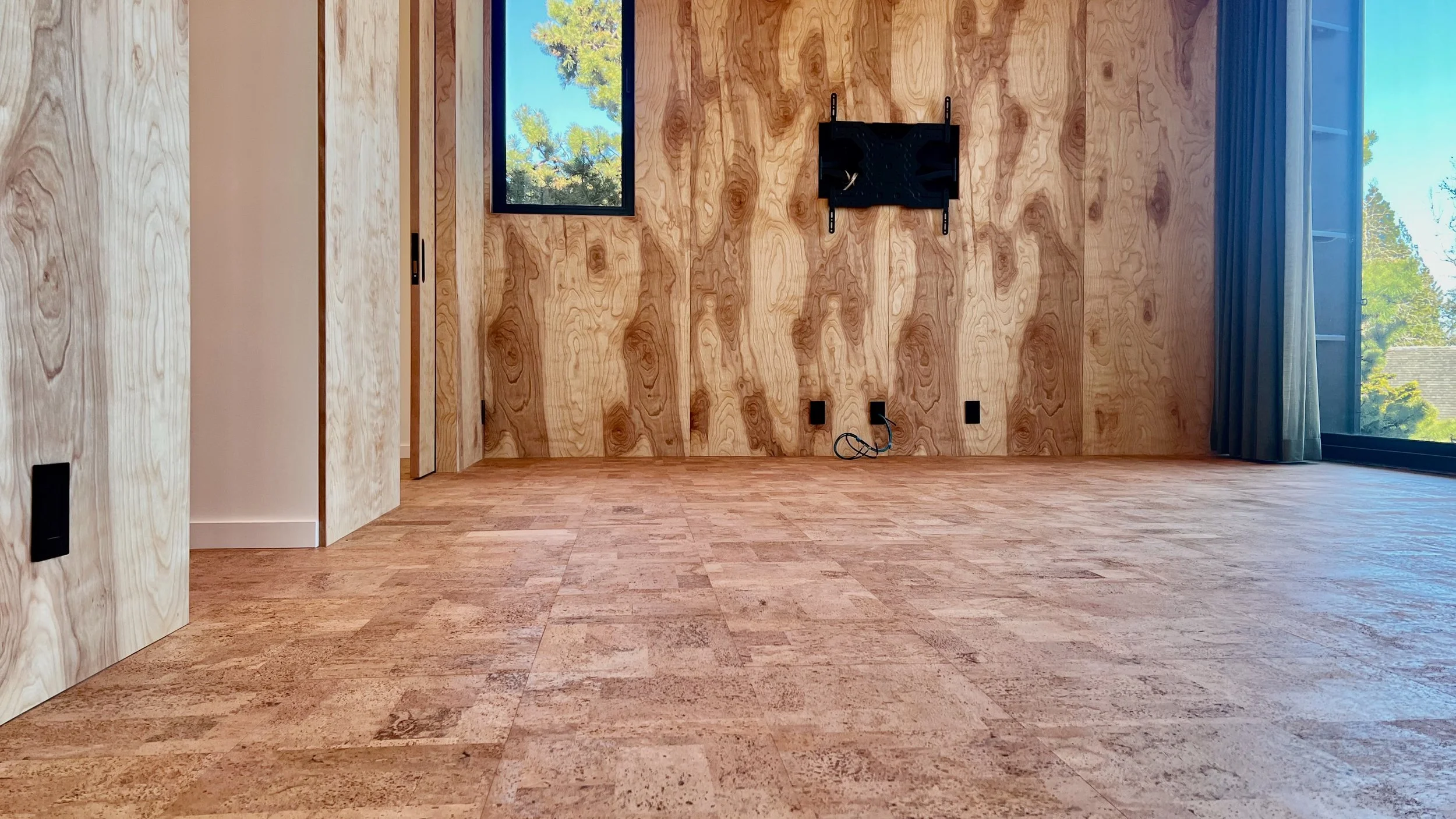 Empty room with wood-paneled walls, hardwood floors, a small window showing trees outside, and a wall mount for a TV or monitor.