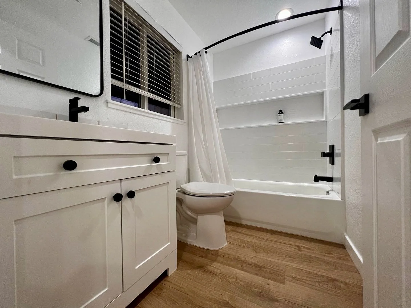bright white restroom with laminate flooring, a subway tile style acrylic tub, and black fixtures. 