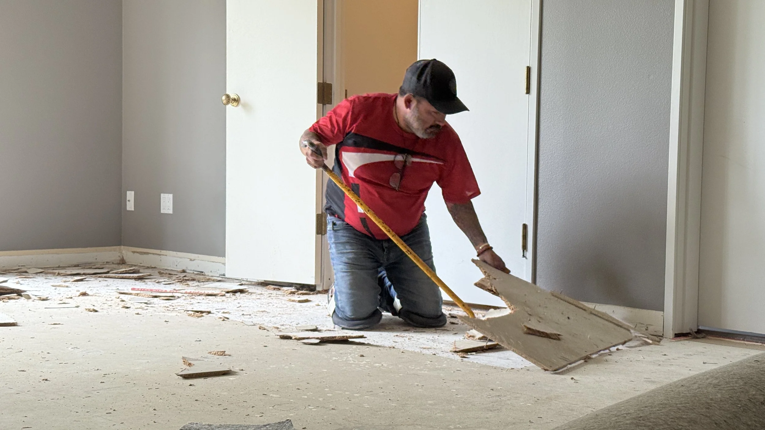 A man wearing a red shirt, black cap, and jeans kneeling on the floor, removing drywall debris with a tool, in a room with gray walls and a door in the background.