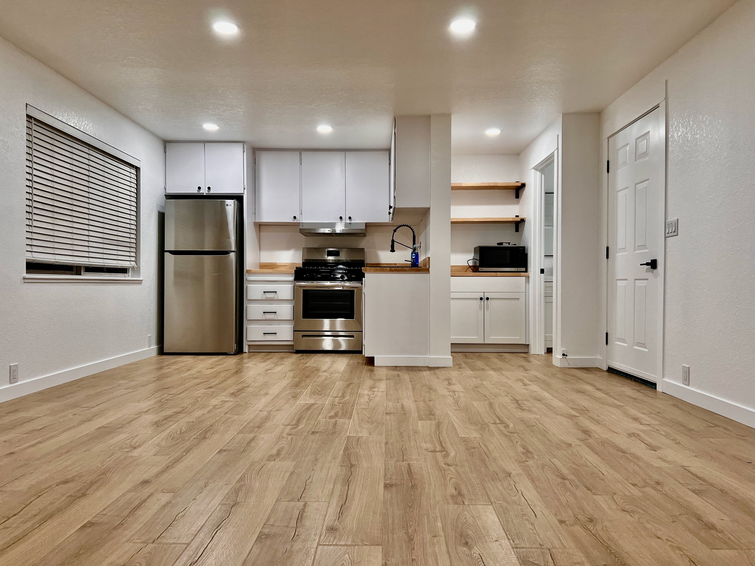 View of a kitchen with white cabinets, black hardware, a stainless steel refrigerator, oven, microwave, white walls, a window with blinds, wooden shelves, and a wooden floor.