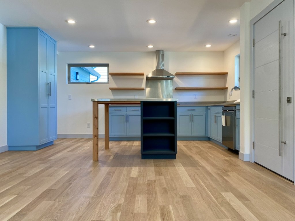 Modern kitchen with light blue cabinets, a small navy blue island with open shelves, wood floating shelves, hardwood flooring, and a stainless steel range hood.