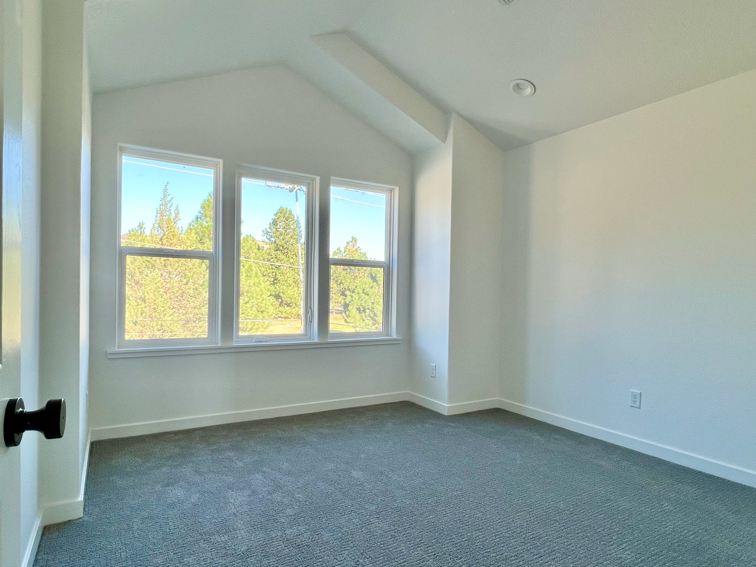 Empty room with large windows, white walls, ceiling, gray carpeted floor, and a partially visible door handle on the left side.