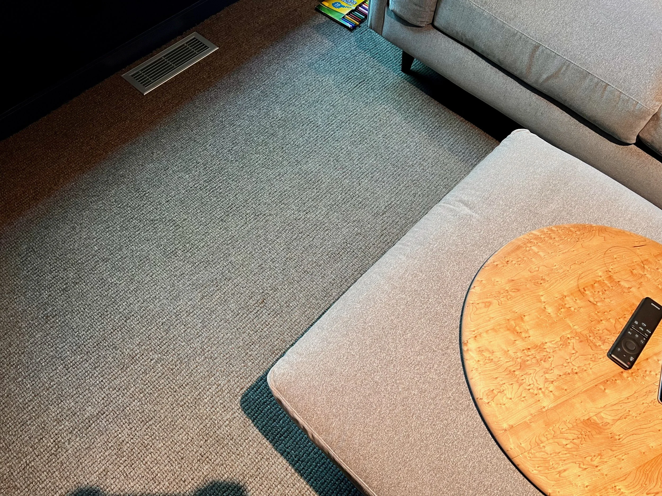 A living room corner with a gray sofa, a round wooden table with a remote control, a box of colored markers on the sofa arm, and a gray and black carpeted floor with an air vent.