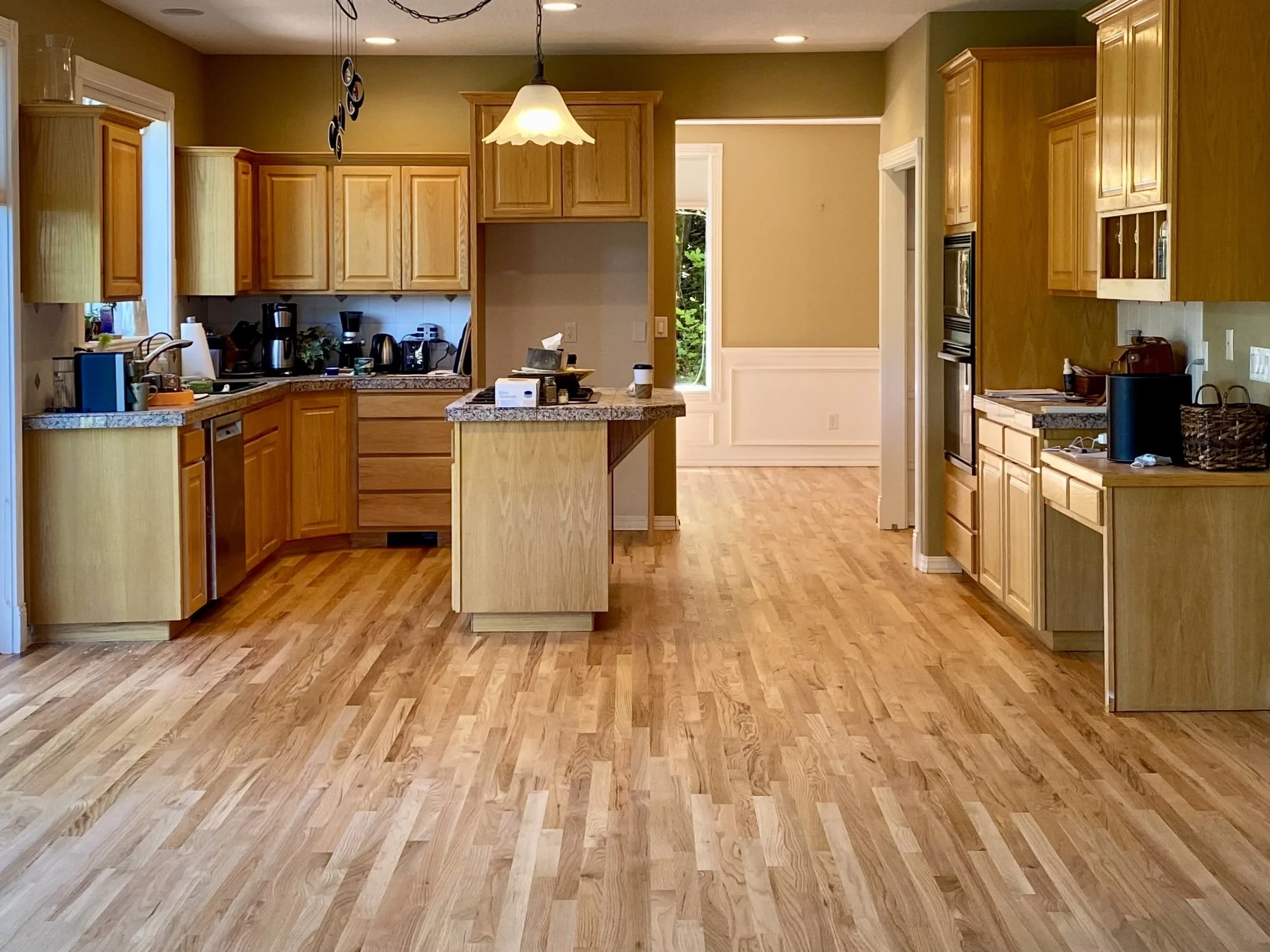A spacious kitchen with wooden cabinets, hardwood flooring, and an island in the center. There are various kitchen appliances and items on the countertops, with natural light coming through the windows and a doorway leading outside.