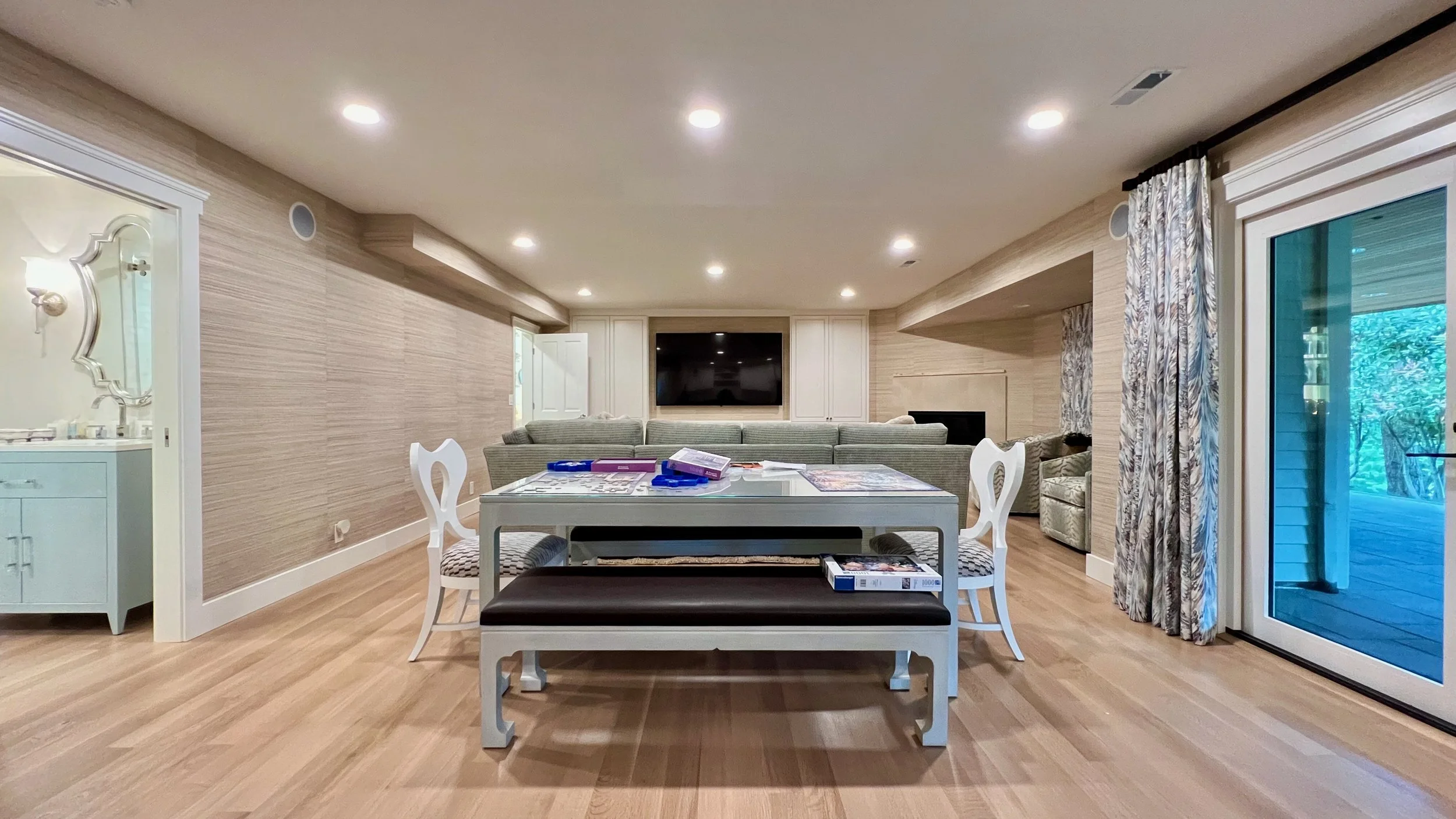 Living room with a dining table, chairs, a sectional sofa, and a wall-mounted TV, with sliding glass door revealing outdoor patio and garden.