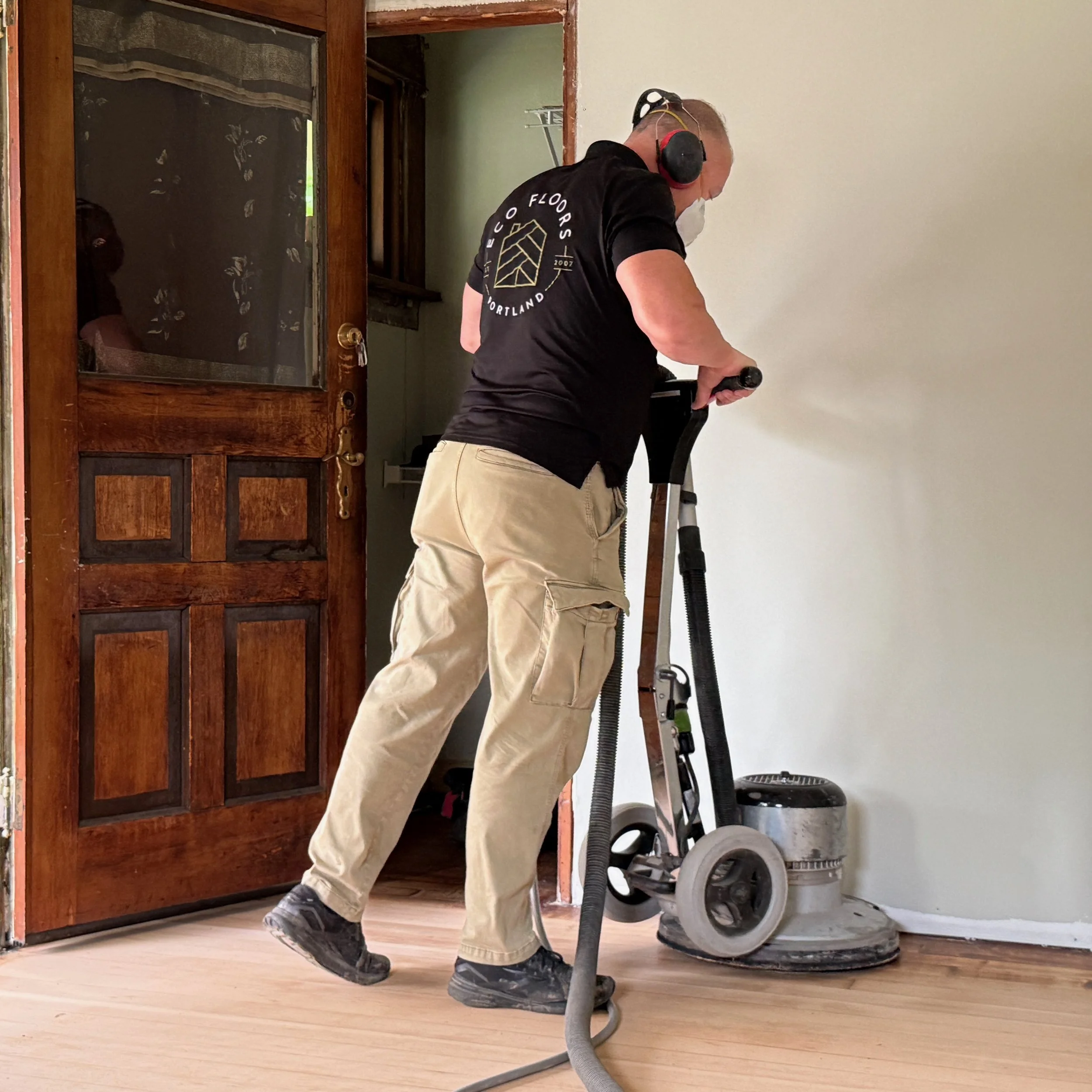 A man wearing a black T-shirt, beige cargo pants, and black shoes is operating a floor sander in a room with white walls and wooden flooring. He is wearing protective ear muffs and a dust mask. There is a wooden cabinet with a glass door on the left, and a vacuum cleaner connected to the floor sander on the right.
