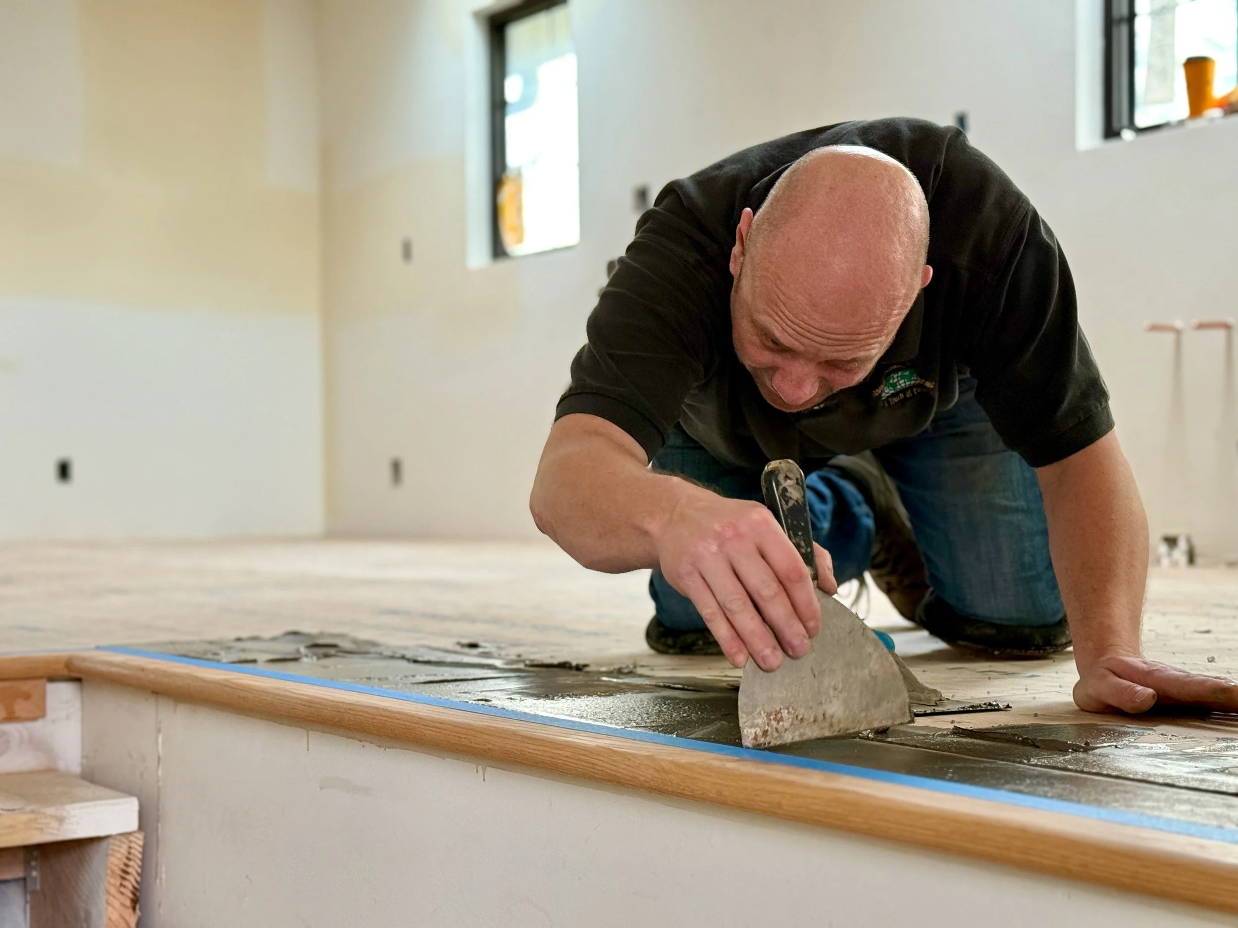 A man with a bald head working on a construction project, kneeling on the floor and smoothing concrete with a trowel inside a partially finished building with unfinished walls and windows in the background.