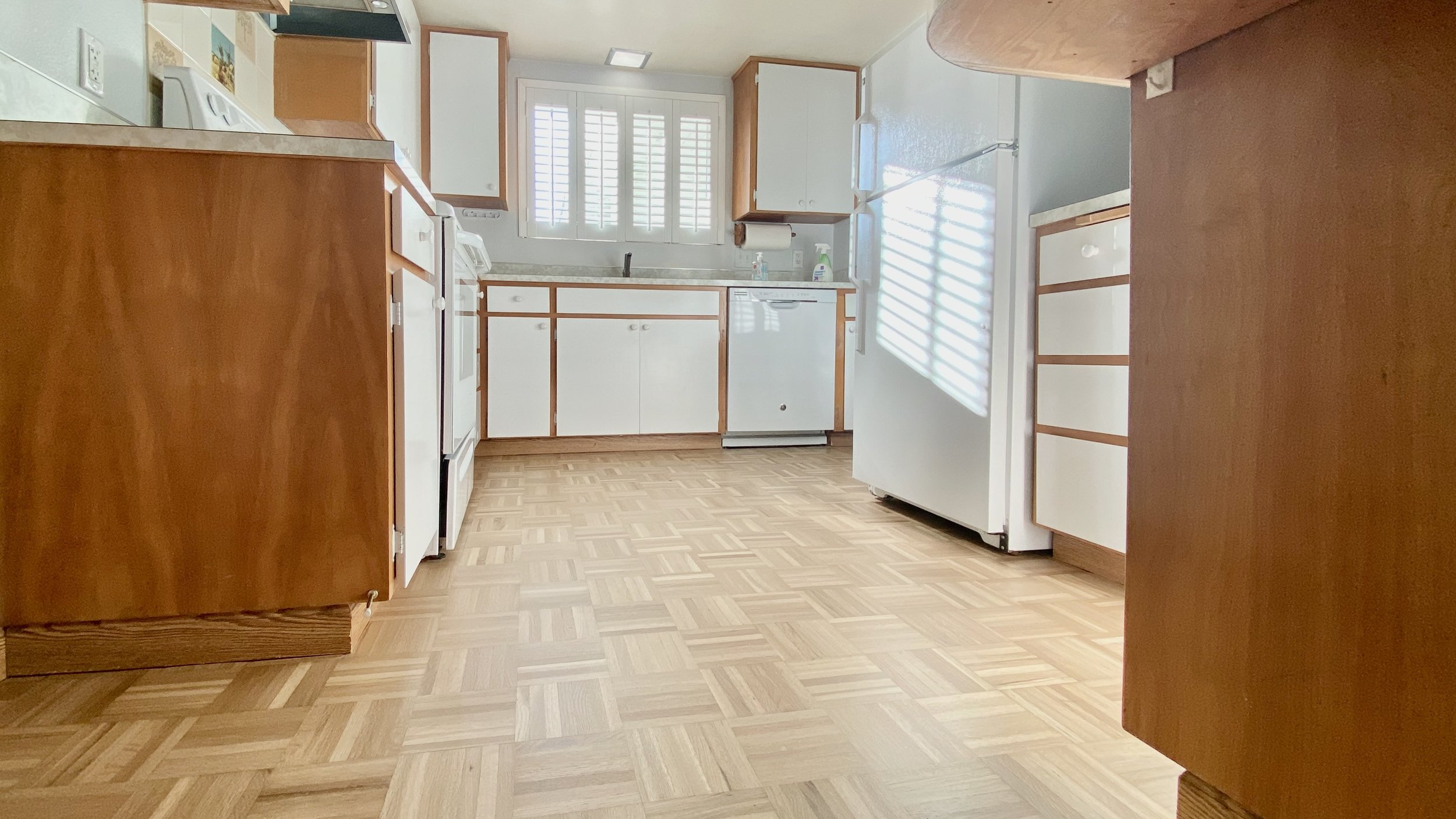 A kitchen with wooden cabinets, white appliances including a refrigerator and dishwasher, a window with white shutters, and a patterned wood floor.