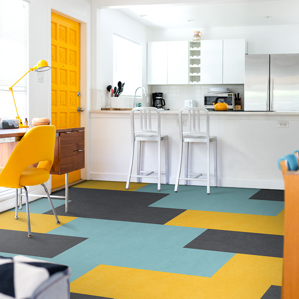 Bright kitchen with a yellow door, white cabinets, and colorful geometric flooring. Yellow chair with a wooden desk, and kitchen appliances including a toaster, microwave, coffee maker, and refrigerator visible.