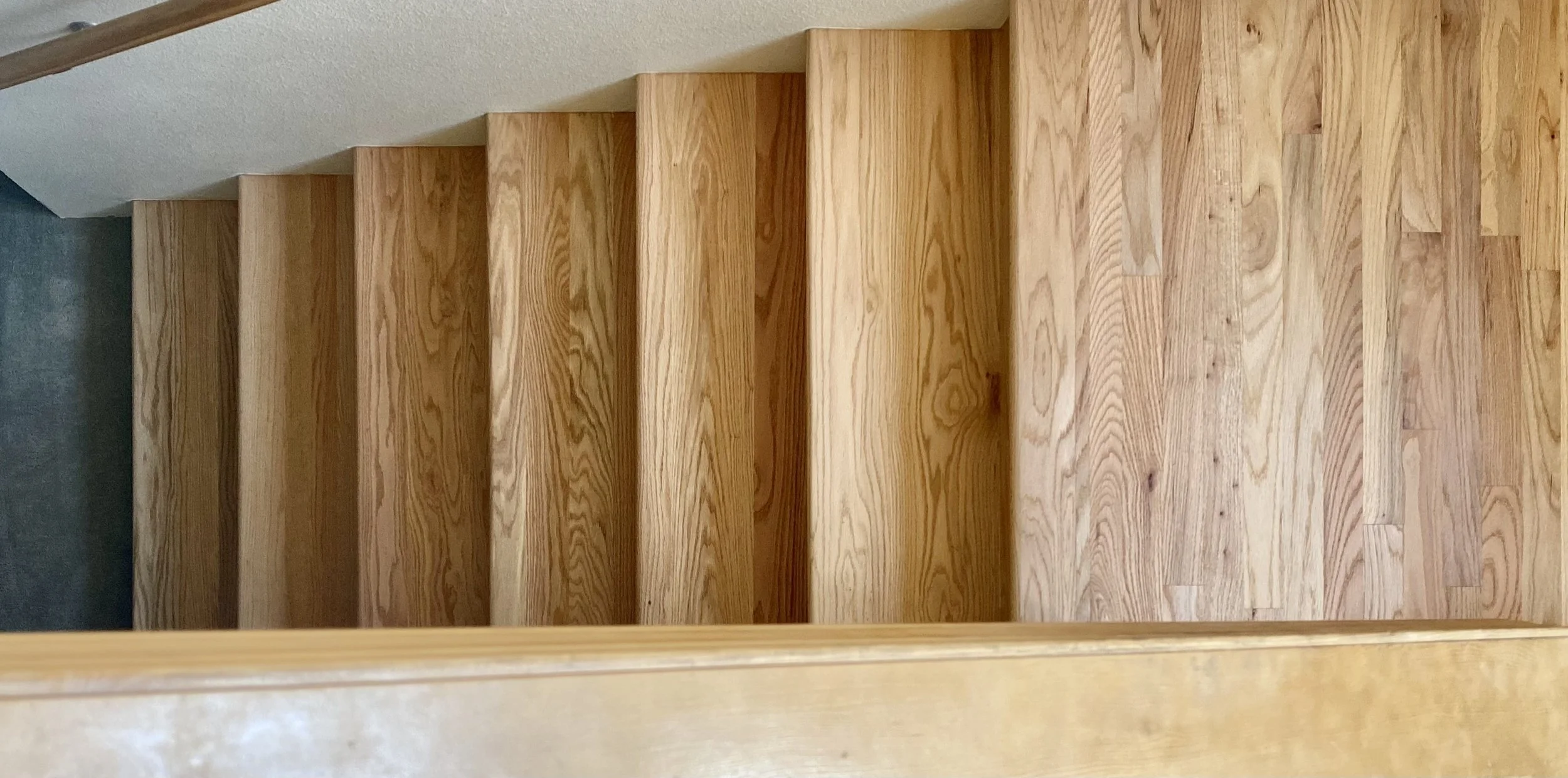 Wooden staircase viewed from above, with light-colored wooden steps and a beige wall on the right side.