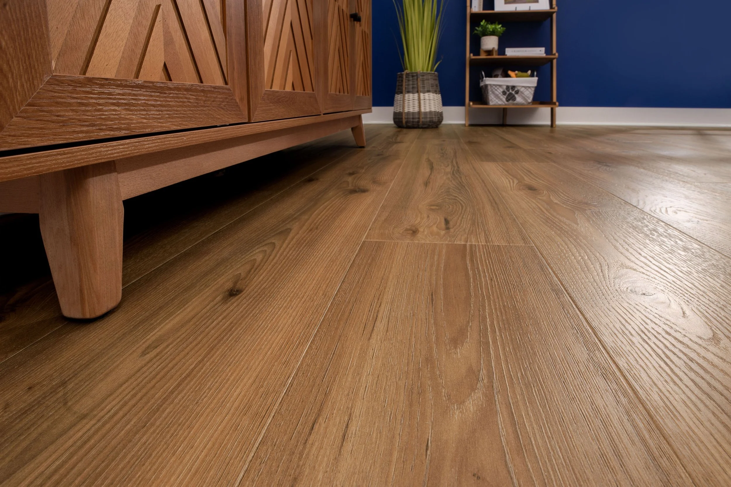 Close-up of a wooden furniture piece and hardwood floor in a room with blue walls, a potted plant, and bookshelf in the background.