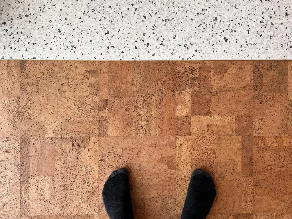 A person's feet on a reddish-brown cork floor next to a section of speckled white terrazzo.