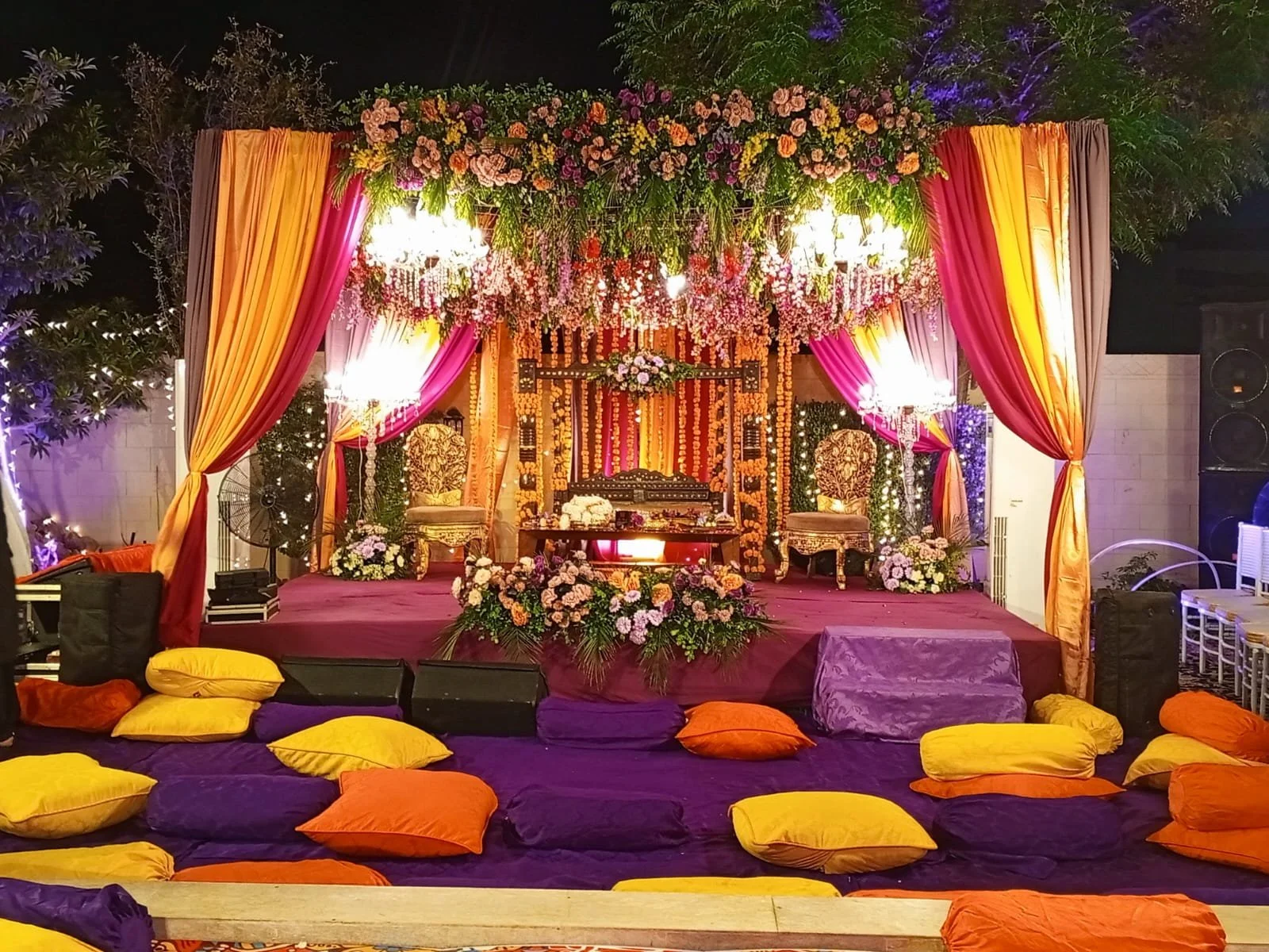 Decorated stage with pink, orange, and purple drapes, floral arrangements, and seating for a celebration, likely a wedding or event.