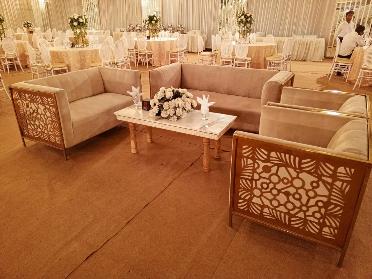 Elegant wedding reception hall with round tables, white chairs, floral centerpieces, and a comfortable seating area with beige sofas around a marble-topped coffee table.