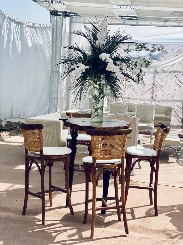 A tall vase with white flowers and dark green leaves, placed on a round wooden table, set in a bright room with white curtains and antique-style chairs and sofas.