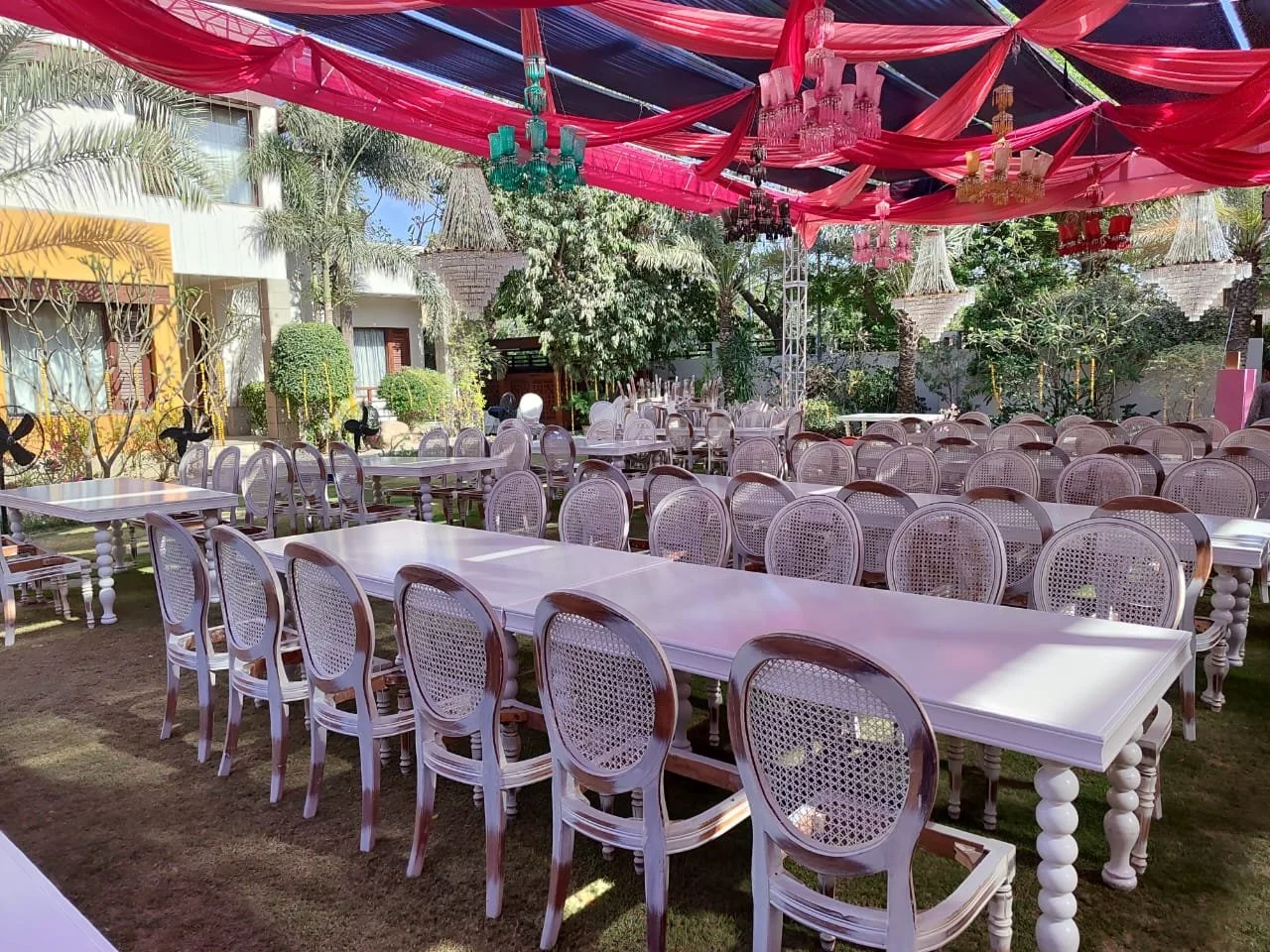Outdoor event space decorated with pink and blue fabric drapes and chandeliers, featuring long tables and white chairs, surrounded by trees and tropical plants.