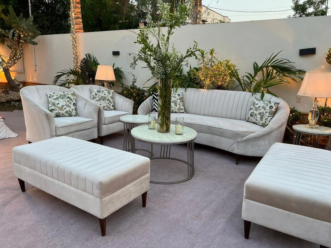 Outdoor patio with white upholstered furniture, including a sofa, two armchairs, and two ottomans. There are marble-topped tables, large glass vases with flowers, and lamps providing warm lighting. Green plants and trees are visible along a white wal