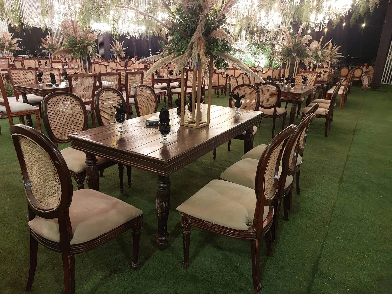 Elegant outdoor dining area with wooden tables and chairs, decorated with black napkins tied into bows, and adorned with floral centerpieces with pampas grass, under hanging string lights on a grassy floor.
