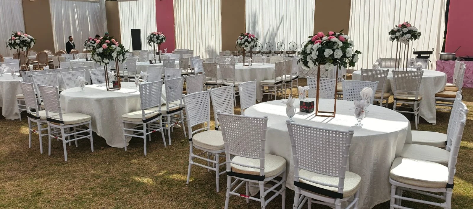 Round banquet tables with white tablecloths and white chairs, decorated with tall floral centerpieces of pink and white flowers, set up for an event with napkins, glasses, and small boxes on the tables.