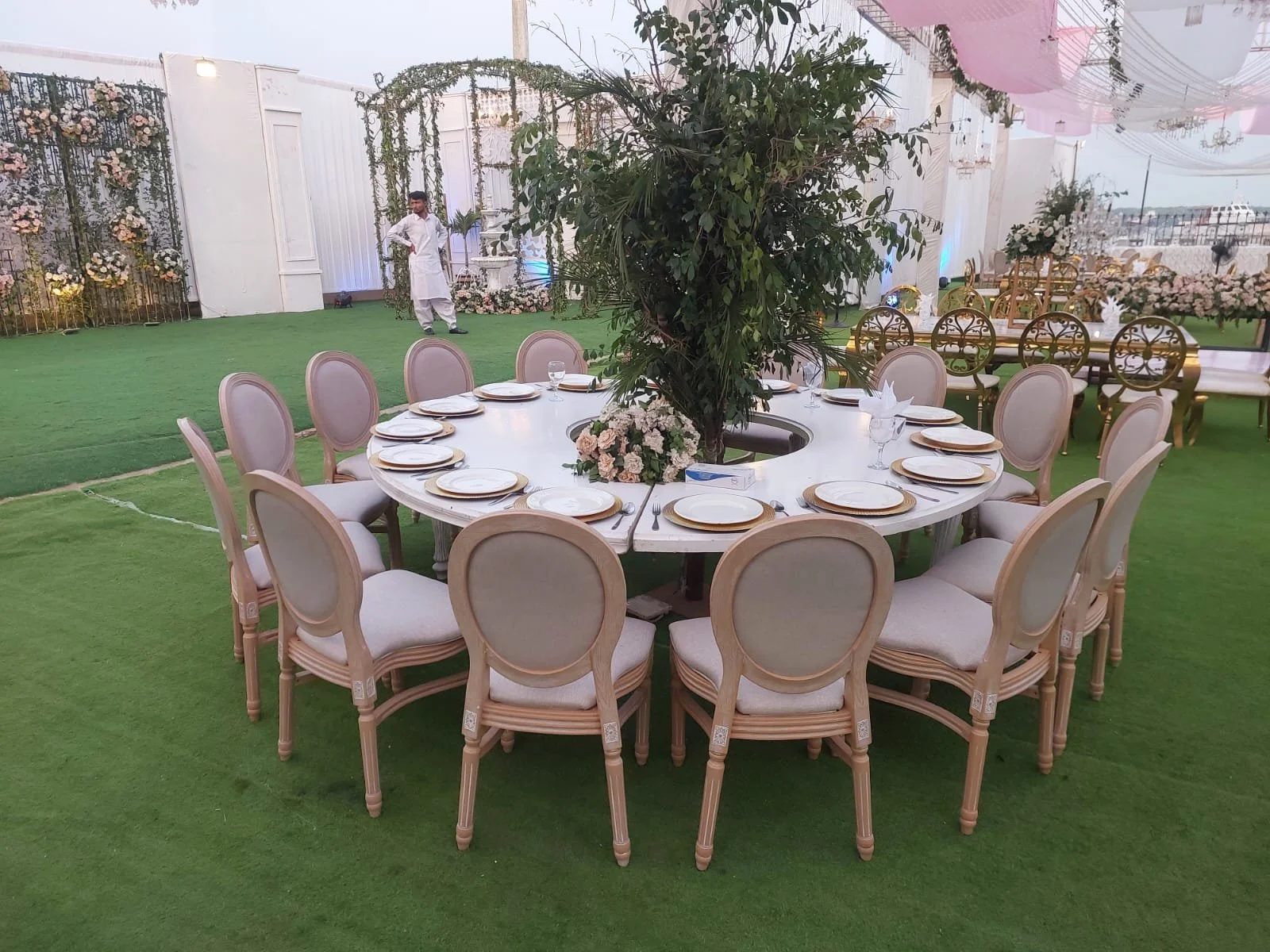 Round banquet table set with white plates, gold-colored chargers, silverware, and glasses, with a large central plant and a floral arrangement, surrounded by cream-colored chairs, outdoors on green grass under a decorated canopy.