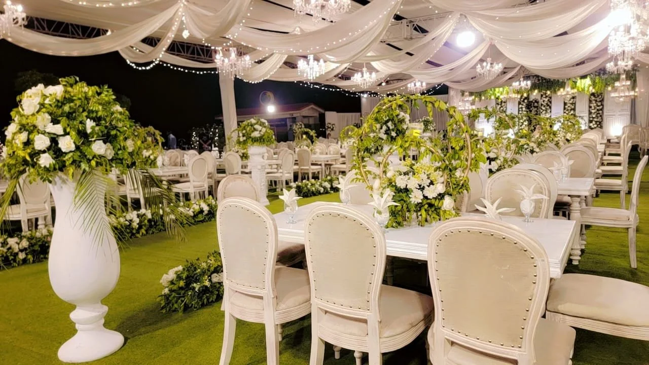 Elegant outdoor wedding reception with white chairs, tables, and floral arrangements in a decorated tent with draped white fabric and chandeliers.