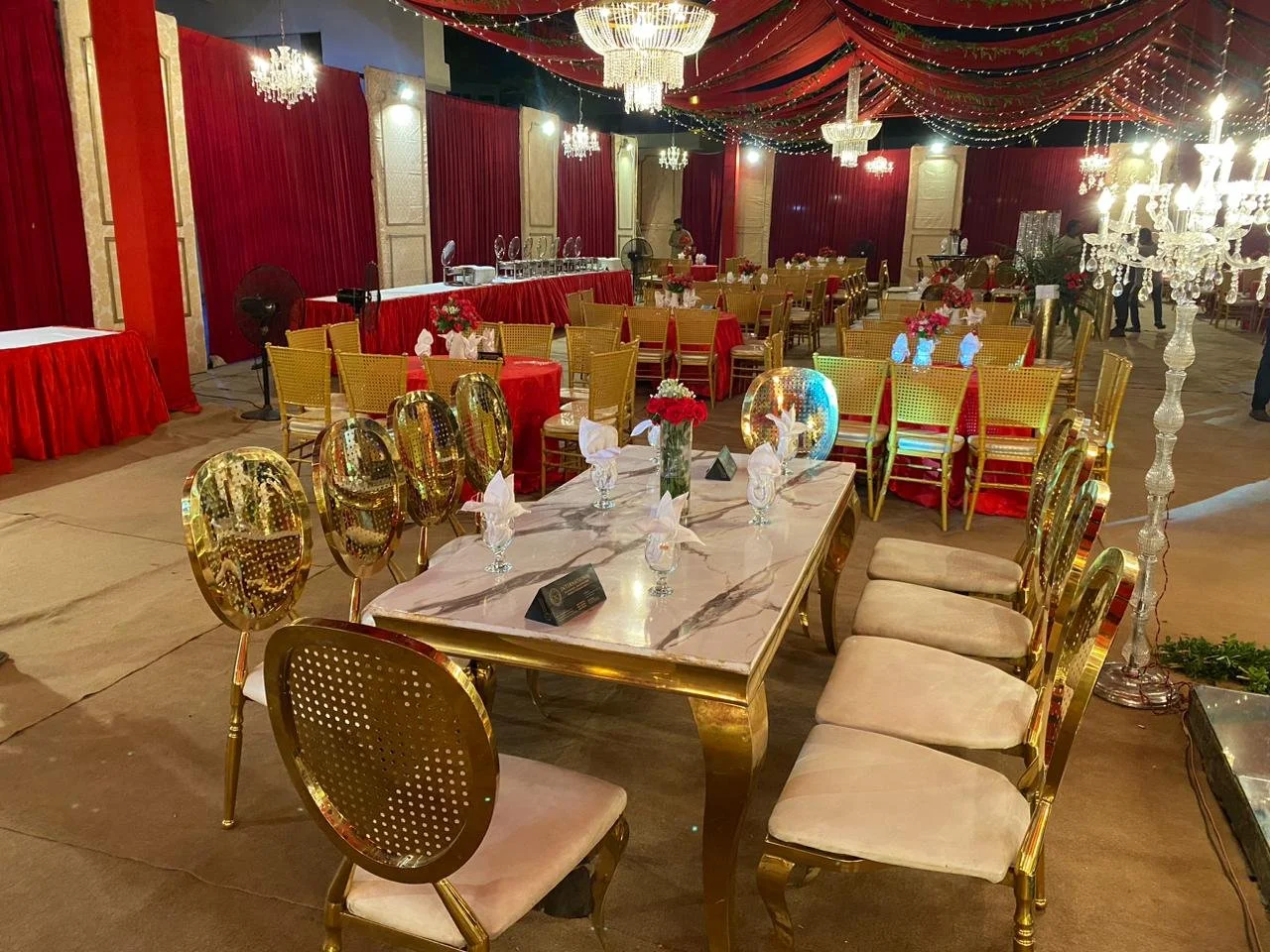 Elegant banquet hall with gold and beige chairs, decorated tables with floral centerpieces, red and gold drapery, chandeliers, and string lights.