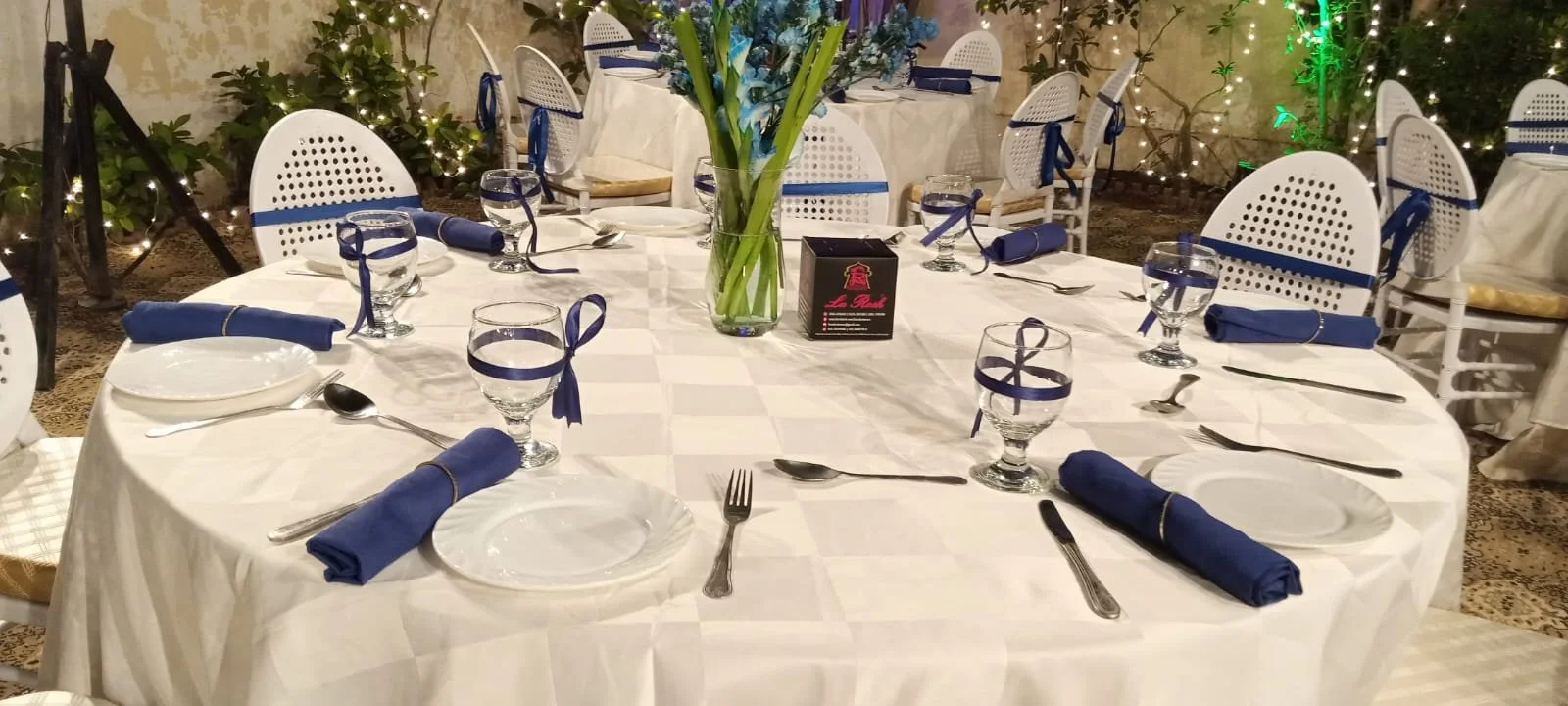 Round banquet table set for a formal dinner with white tablecloth, blue napkins, water glasses with blue ribbons, white plates, and silverware, decorated with a tall floral centerpiece of blue flowers in a glass vase, with a black box and greenery in