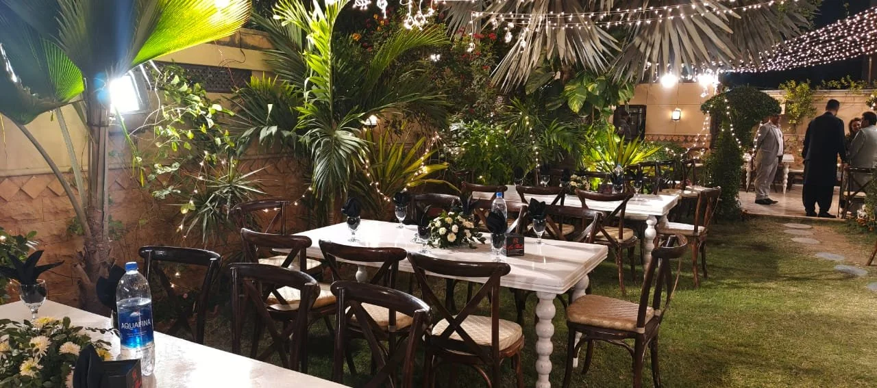 Outdoor dining area at night with decorated tables, white tablecloths, black napkins, and floral centerpieces, surrounded by lush green plants and string lights.