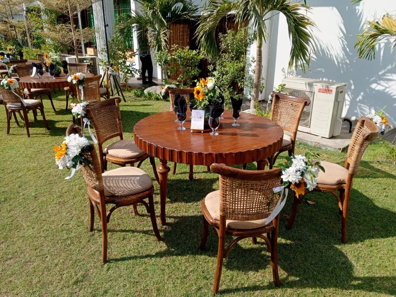 Outdoor table setting with eight chairs decorated with white and yellow flowers on an outdoor lawn, surrounded by trees and plants.