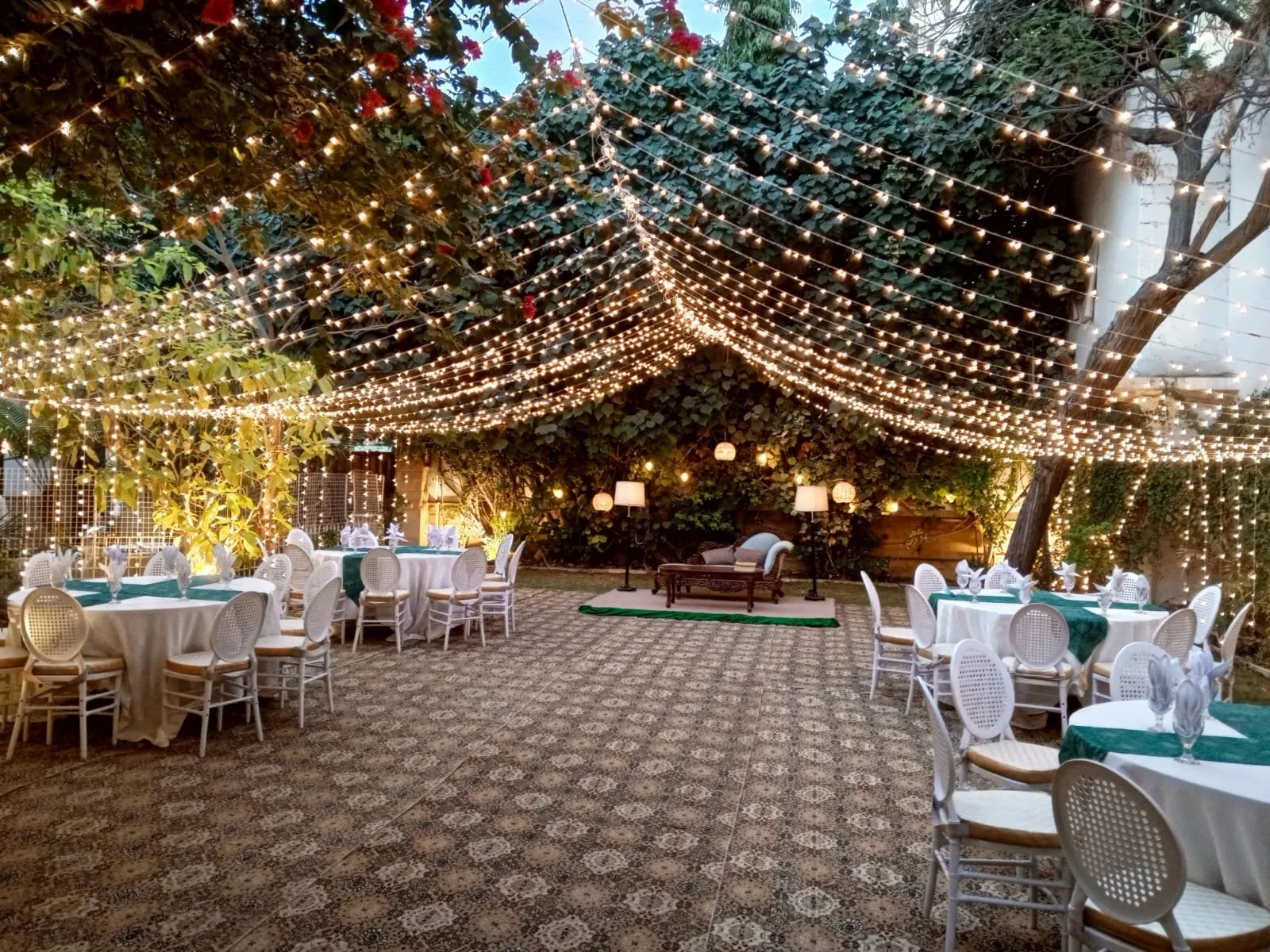 Outdoor event space decorated with string lights hanging from trees, with round tables covered with white tablecloths and green runners, set with napkins and glassware, and a small stage with a bench, lamps, and a backdrop of foliage