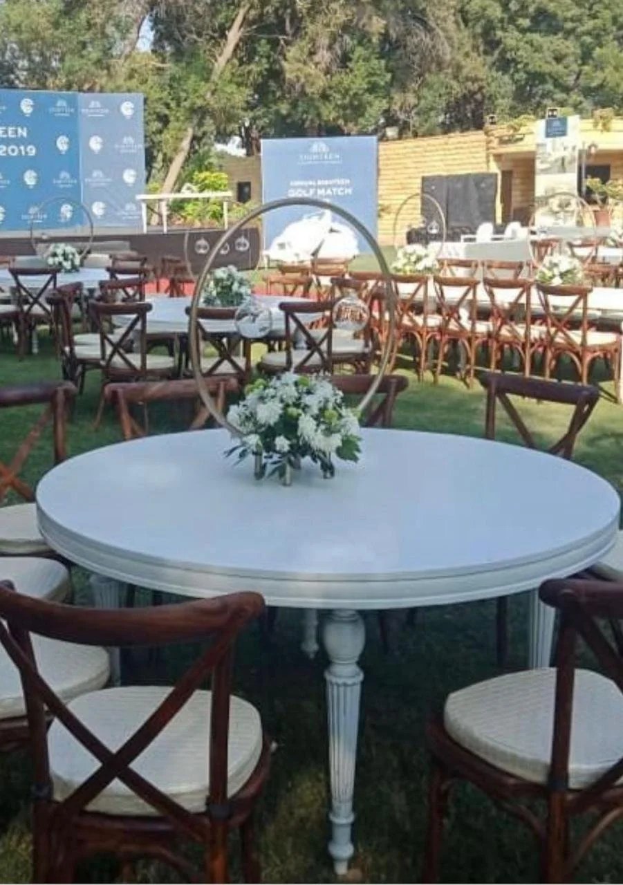 Outdoor event setup with round white tables, wooden chairs, and floral centerpieces, with a blue backdrop and banners.