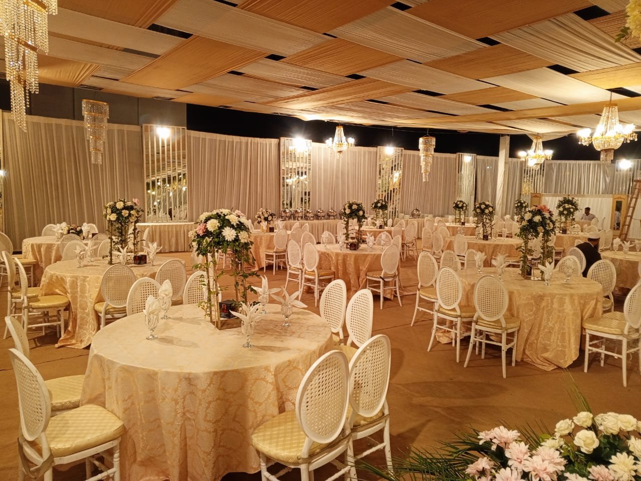 Elegant banquet hall decorated with round tables covered in gold tablecloths, adorned with white floral centerpieces, and surrounded by white chairs with yellow cushions. The ceiling has a decorative wooden pattern with chandeliers hanging down. The 