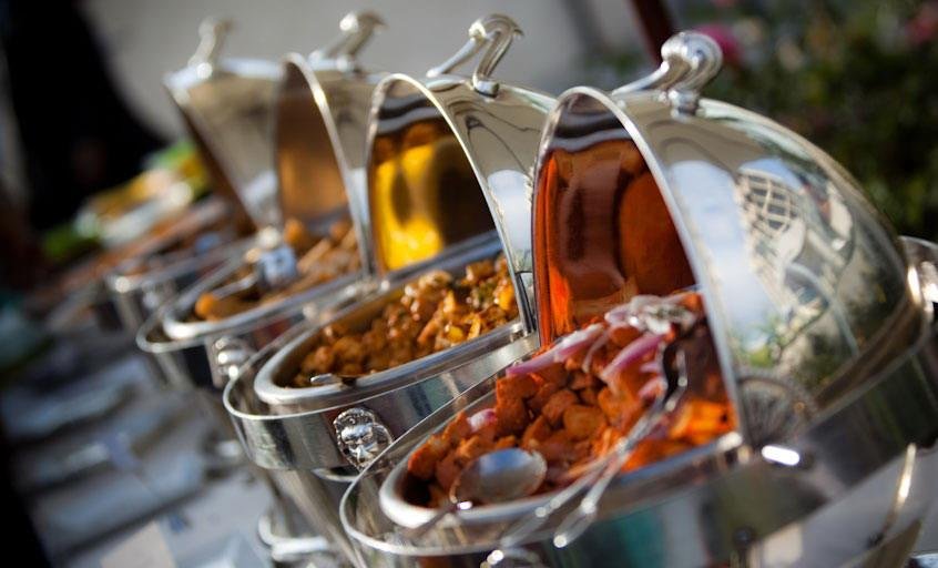 Various hot food dishes in stainless steel chafing dishes with lids open, at a buffet.
