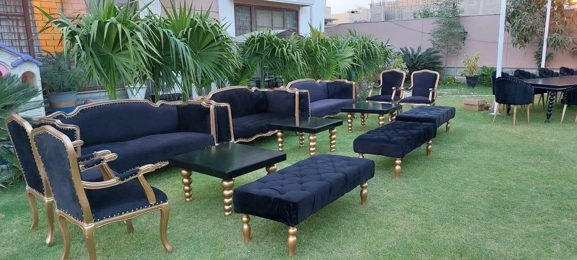 Outdoor garden seating with black velvet sofas, armchairs, and ottomans with gold accents, arranged on a grassy lawn with green plants and trees in the background.