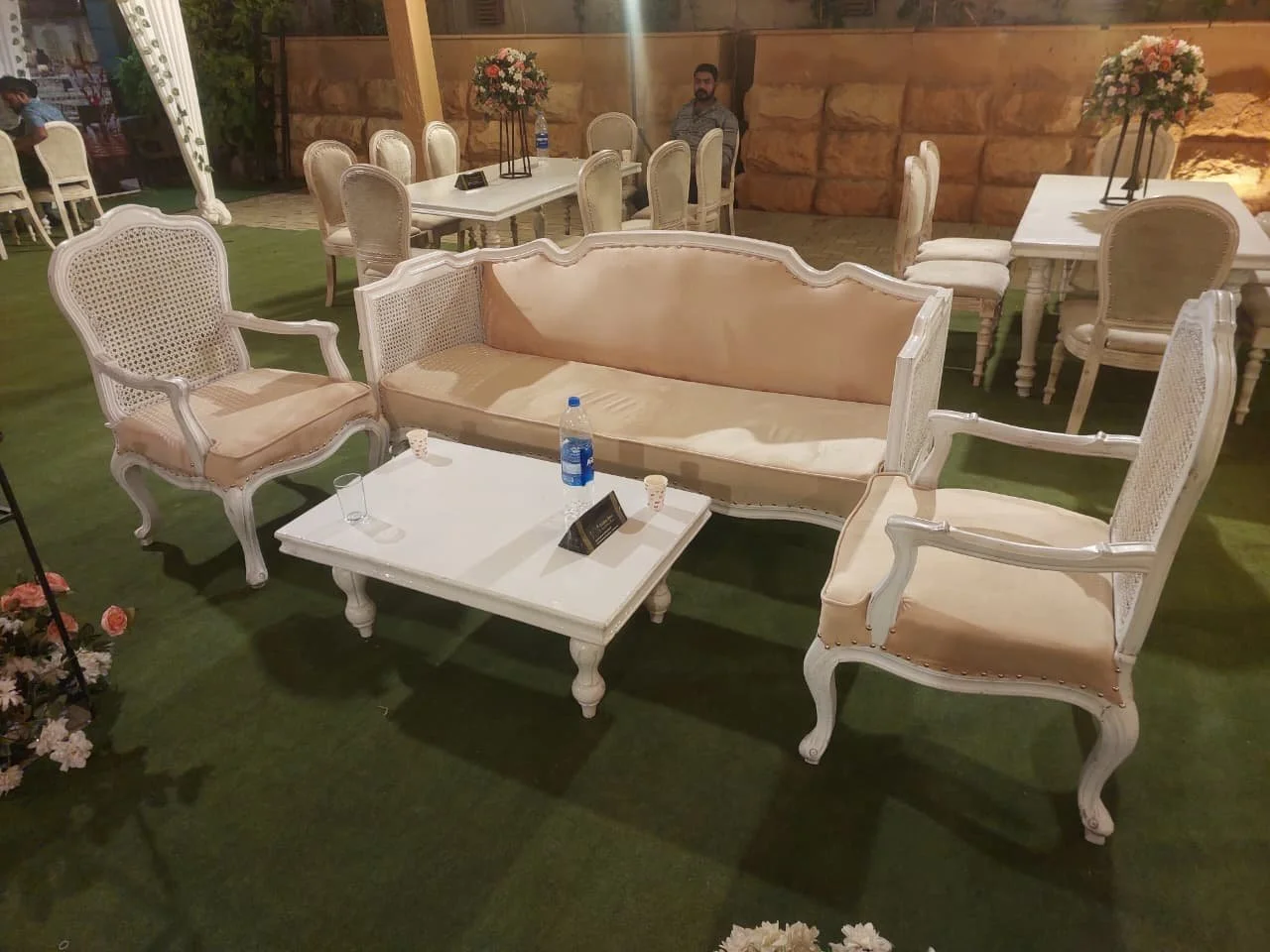 Elegant outdoor seating arrangement with vintage white upholstered chairs, a matching loveseat, and a coffee table, decorated with flower arrangements in vases.