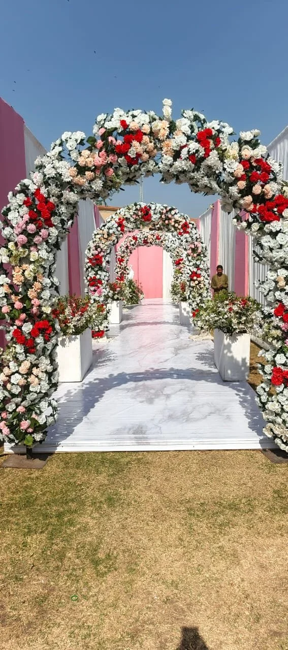 A pathway decorated with multiple archways made of colorful flowers, including white, pink, red, and peach, leading to a pink backdrop, set up outdoors on a sunny day.