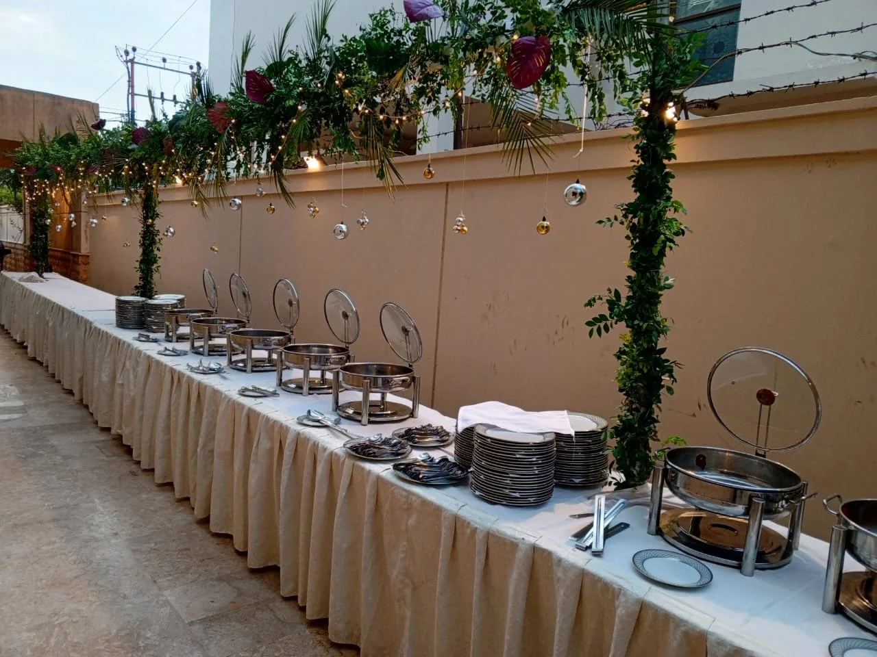 Buffet table set up outdoors with chafing dishes, plates, and utensils. Decorated with greenery, string lights, and hanging ornaments.