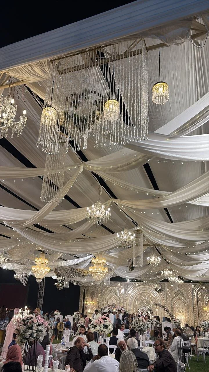 Elegant indoor wedding reception with white draped fabric ceilings, multiple chandeliers, and hanging crystal chandeliers, decorated with floral arrangements, with guests seated at tables and a decorated main stage in the background.