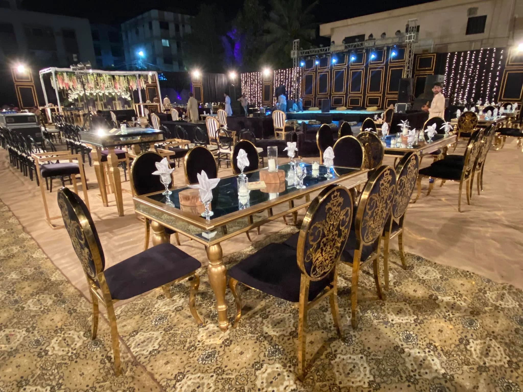 Elegant outdoor event setup with multiple decorated tables, ornate chairs, and a stage in the background, illuminated by string lights and spotlights at night.