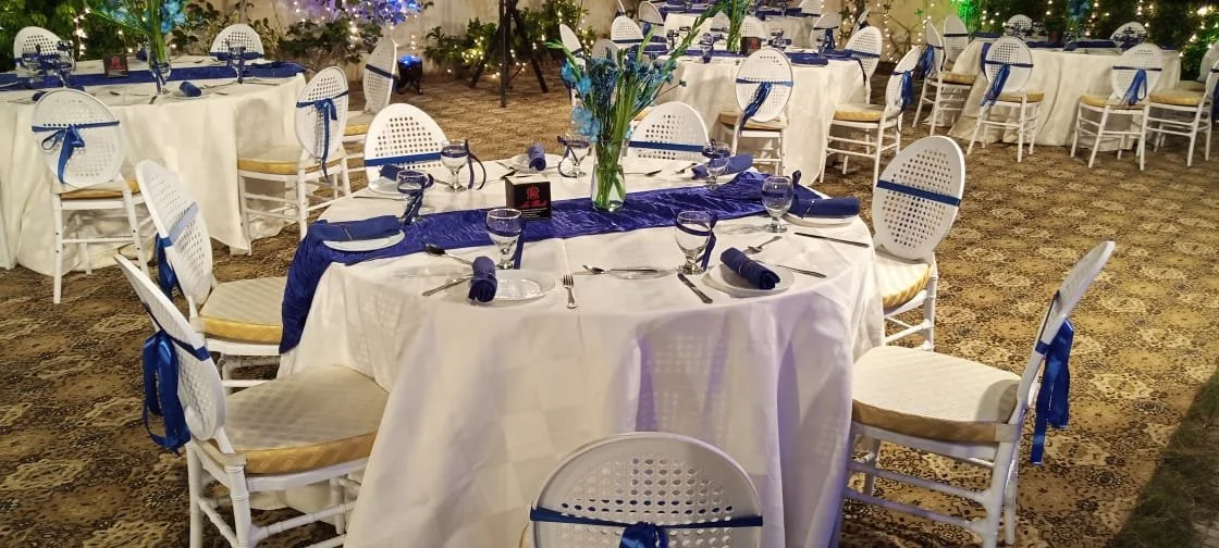 Round banquet tables with white tablecloths, blue napkins, and glassware, decorated with blue flowers in the center, set for an event or party, in an outdoor setting with string lights.