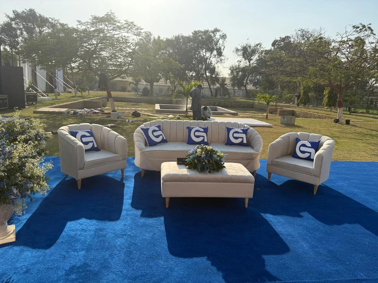 Outdoor seating area with white sofas and armchairs on a blue carpet, decorative pillows with a white G logo, a flower arrangement on a matching ottoman, green trees, and two people working in the background.