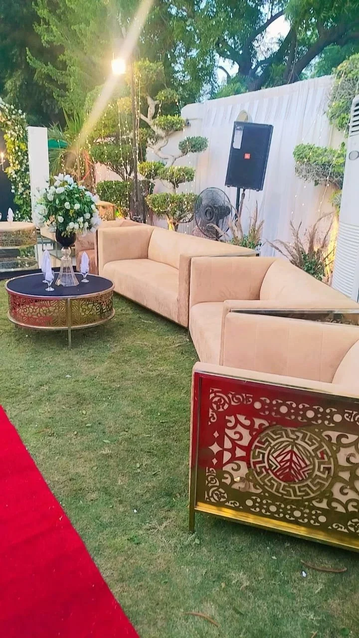 Outdoor event setup with beige sofas, a round black coffee table, and floral arrangements, surrounded by greenery and decorative fencing.