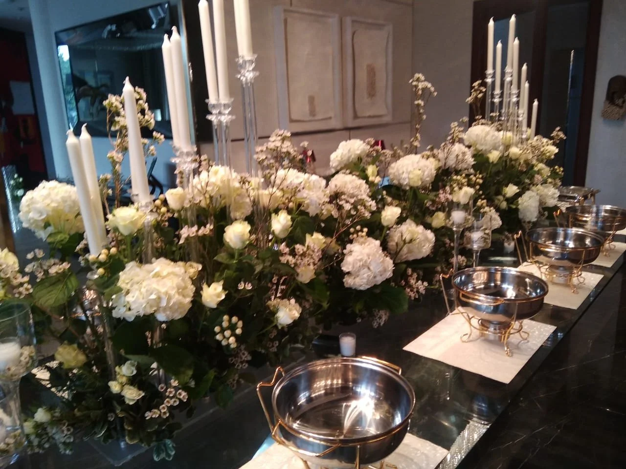 A table decorated with white floral arrangements, tall white candles in glass holders, and empty silver bowls on white placemats.