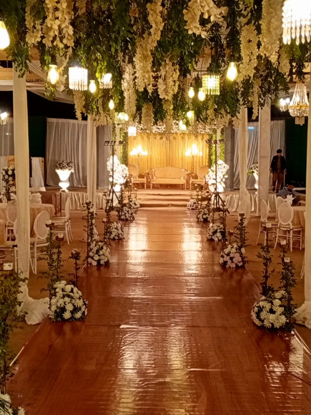 Wedding or event venue decorated with hanging white flowers, green foliage, and warm lighting, featuring a wooden aisle leading to a stage with golden seating and illuminated backdrop.