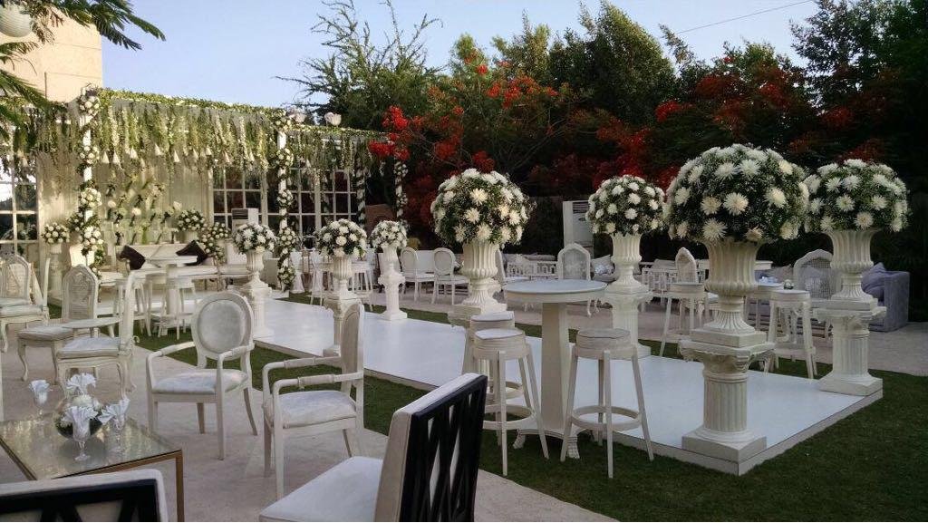 Elegant outdoor event setup with white chairs, tables, and large floral arrangements in white vases, decorated with white flowers, set on a white dance floor with greenery and trees in the background.