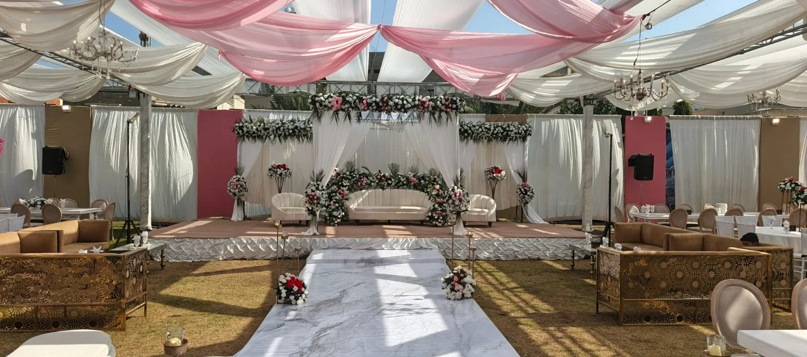 A decorated outdoor wedding stage with white and pink drapes, floral arrangements, and seating, set up on a grassy area.