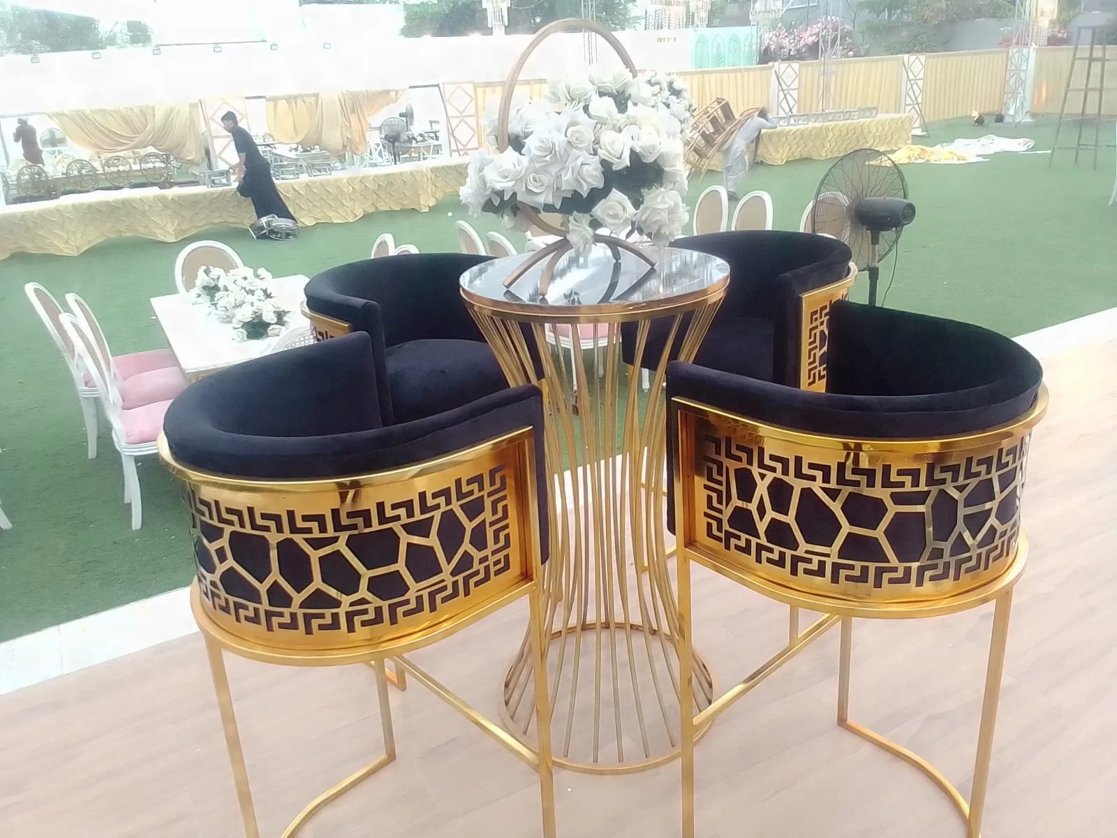 A round table with a floral centerpiece and four black velvet armchairs with gold geometric patterned backs, set on a wooden floor overlooking a decorated event space with tables, chairs, and ongoing setup work.