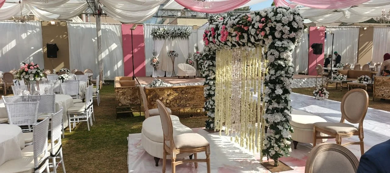 An outdoor wedding reception decorated with white and pink flowers, adorned chairs, and drapery under a tent.